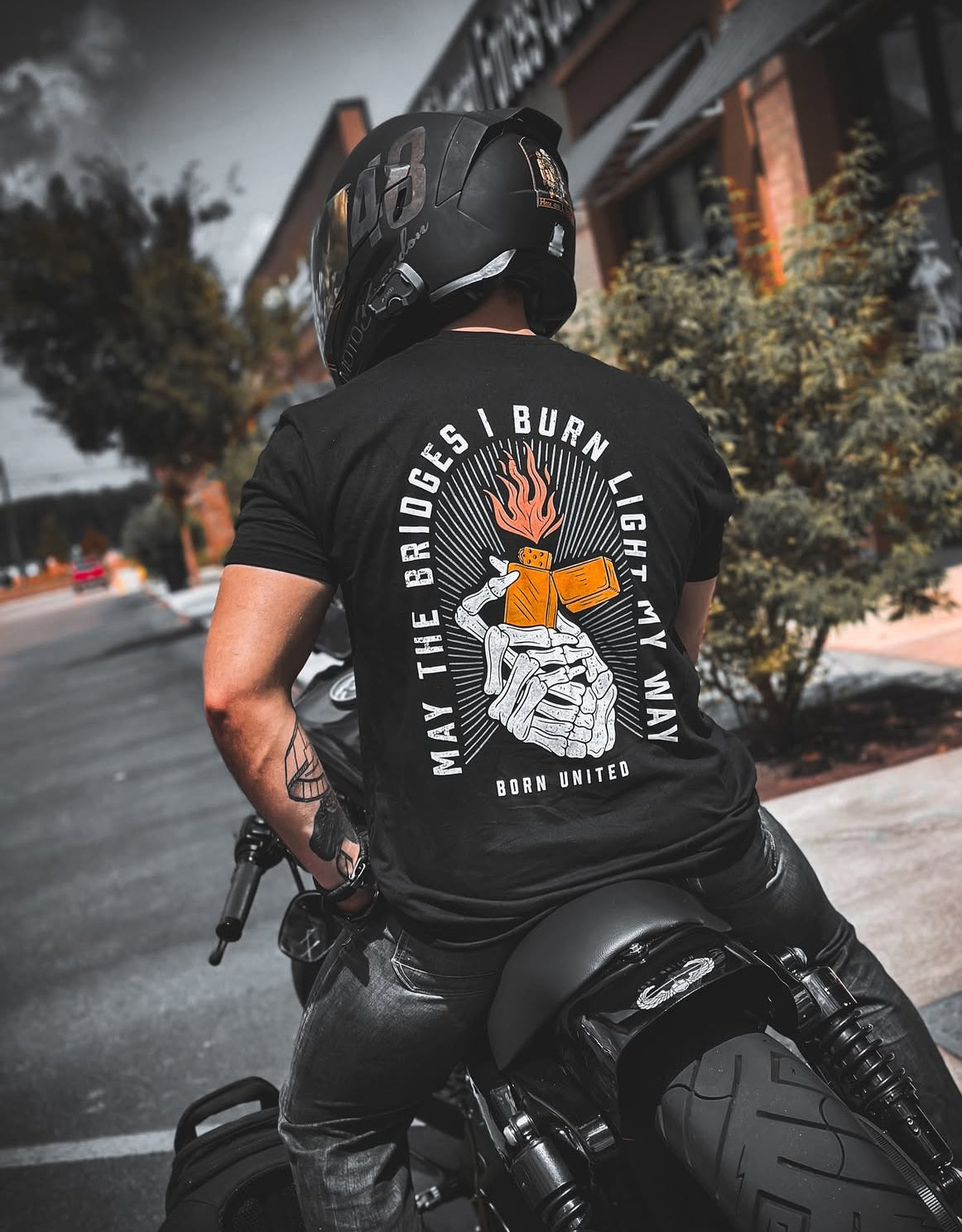 A person in a helmet and a black Burning Bridges (Warehouse) tee by Born United Private Label, featuring skeleton hands shaking over burning dice with the text May the bridges I burn light my way. Born United, rides a motorcycle past a brick building.