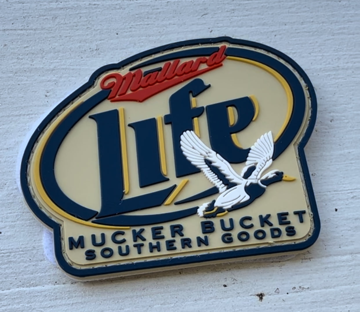 A sign shows Mallard Life with a flying duck illustration, below it reads Mucker Bucket Southern Goods, set against a beige background with blue and red accents.