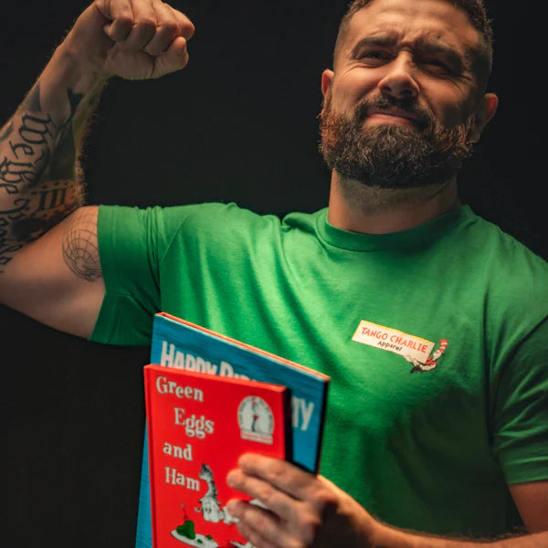 A bearded, tattooed man flexes his bicep to showcase strength while holding Dr. Seuss books: Green Eggs and Ham and Happy Birthday to You! He wears a green St. Seuss Tee from Tango Charlie Apparel with a red name tag on the chest.