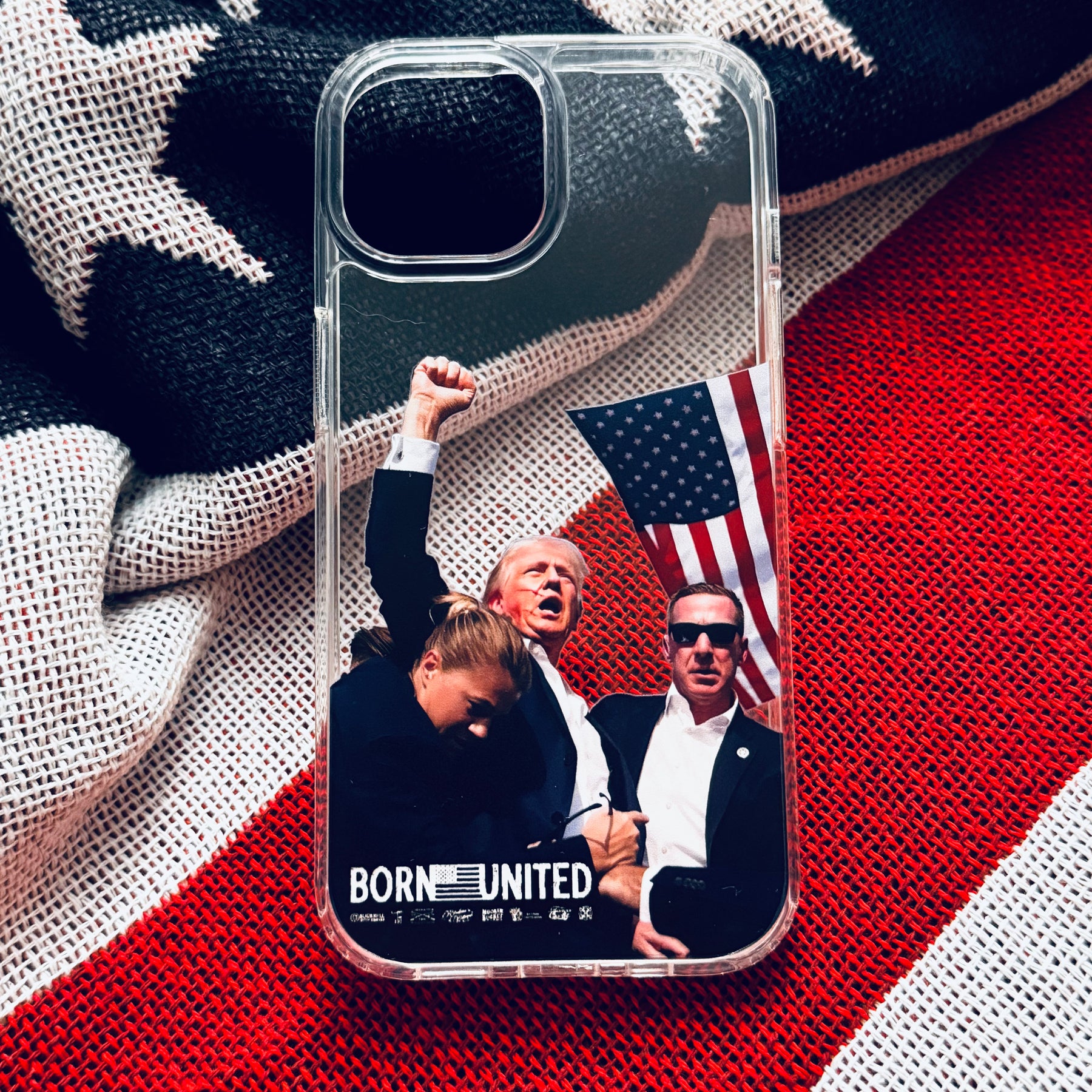 The Forever Legend iPhone Case by Born United Private Label features three men—one raising a fist, another holding an American flag—with “BORN UNITED” below, all set against an American flag fabric background. Transparent design.