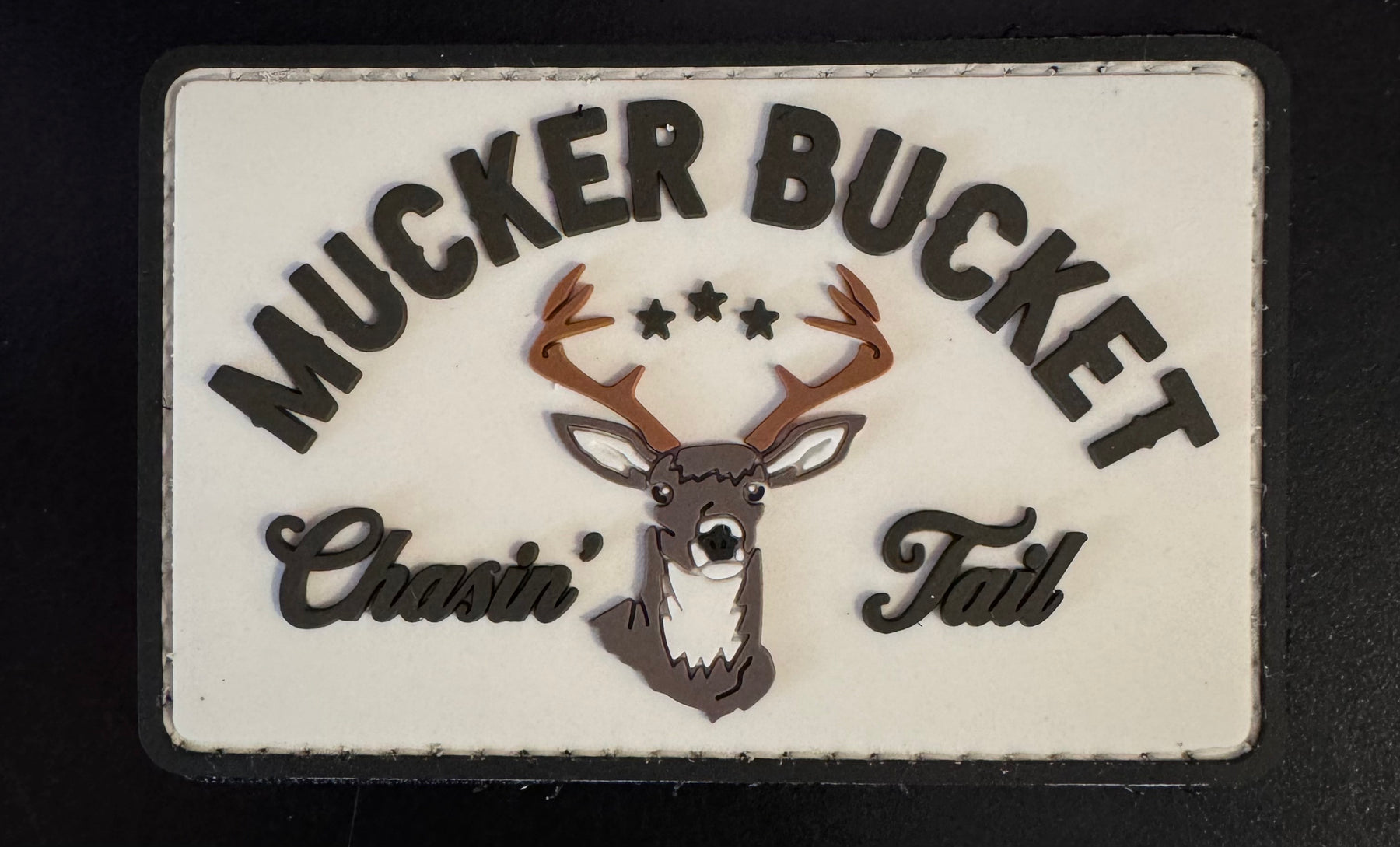 A rectangular sign featuring Mucker Bucket at the top and a deer head with antlers surrounded by three stars in the center, with Chasin Tail beneath it. The background is white, showcasing black text and details matching the Chasin Tail PVC Patch by Mucker Bucket.
