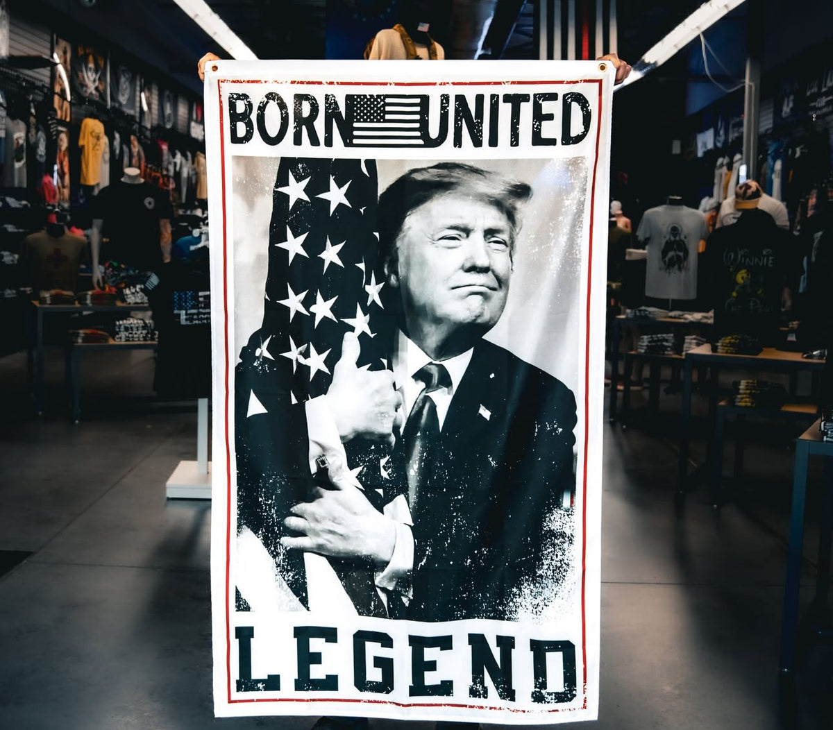 In a clothing store, the Legend Trump Flag by Born United Private Label is displayed, featuring a man with an American flag and BORN UNITED and LEGEND, subtly evoking 2024 election themes and the essence of Make America Great Again.
