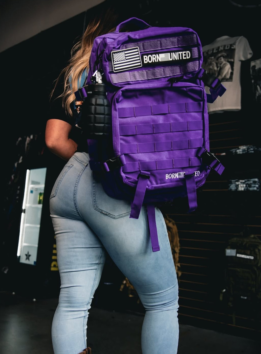A person in light jeans and a black shirt carries a Cosmic Purple Tac-Pac - 45L from Born United Private Label, decorated with a BORN UNITED patch and an American flag. A black water bottle is attached to the side, enhancing the vibrant scene against a backdrop of hanging clothes.