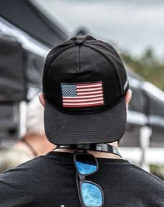 A person is turned away, wearing a Born United Private Label cap featuring an American flag patch. Sunglasses dangle from their shirt collar, paired with a Full Send T-shirt. Black and white striped tents blur in the background.
