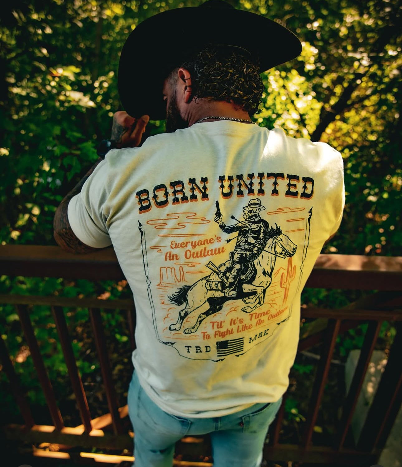 A man in a cowboy hat stands on a wooden deck, wearing the Born United Private Labels Outlaw Tee in tan. The tee features a cowboy on horseback with the phrase Everyones an Outlaw. Lush green foliage provides a fitting backdrop for this rugged scene.