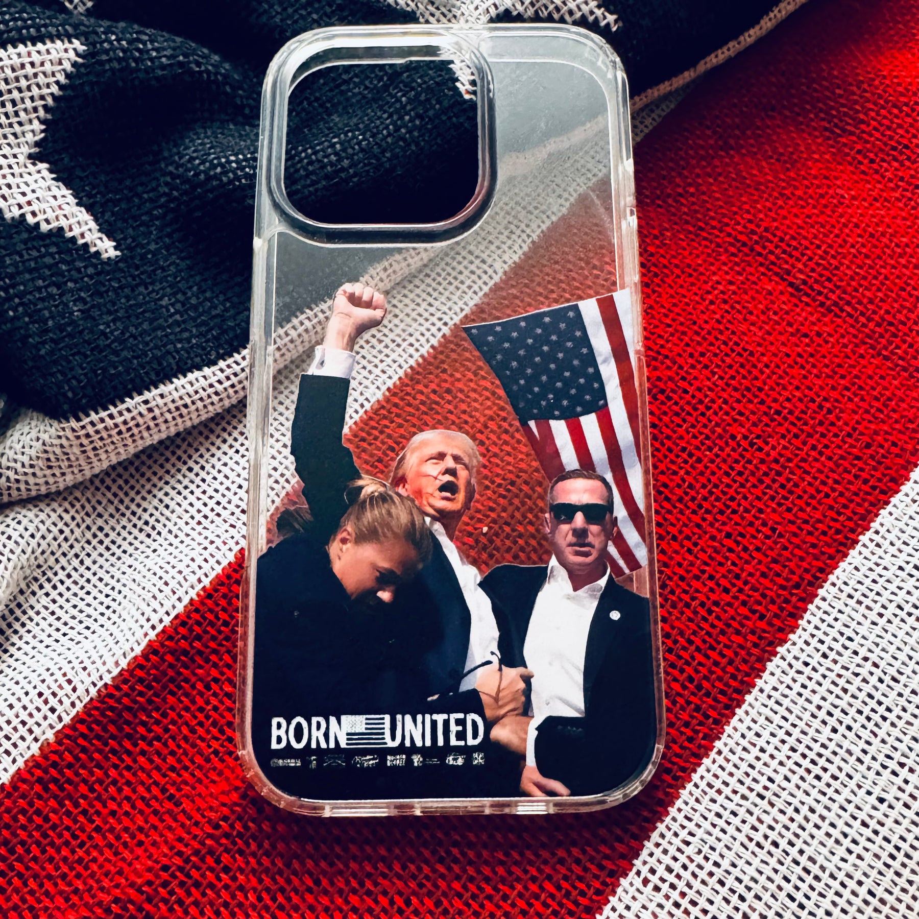 The Forever Legend iPhone Case by Born United Private Label showcases Donald Trump raising his fist, a woman bowing her head, a man in sunglasses, and a U.S. flag, with “BORN UNITED” boldly printed at the bottom.