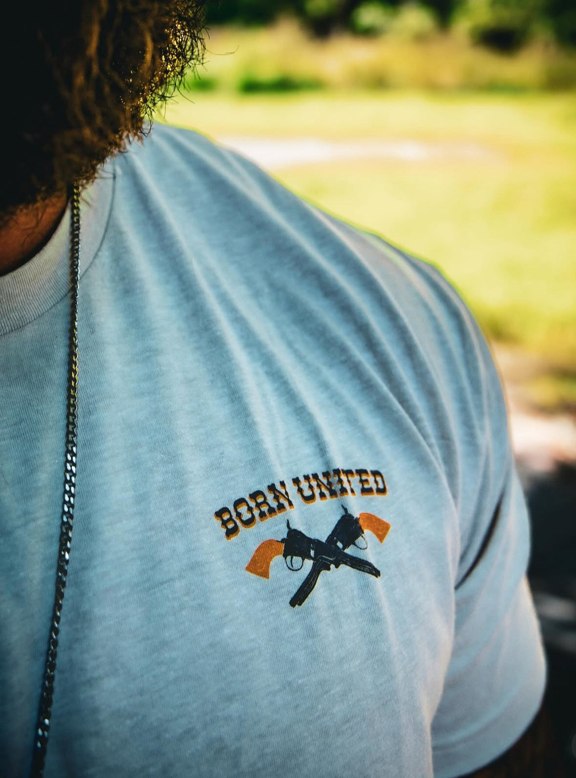 A person is partially visible wearing the Outlaw Tee - Tan by Born United Private Label, featuring a BORN UNITED logo with crossed pistols for an outlaw vibe. A necklace chain can be seen. The blurred background includes grass and trees.