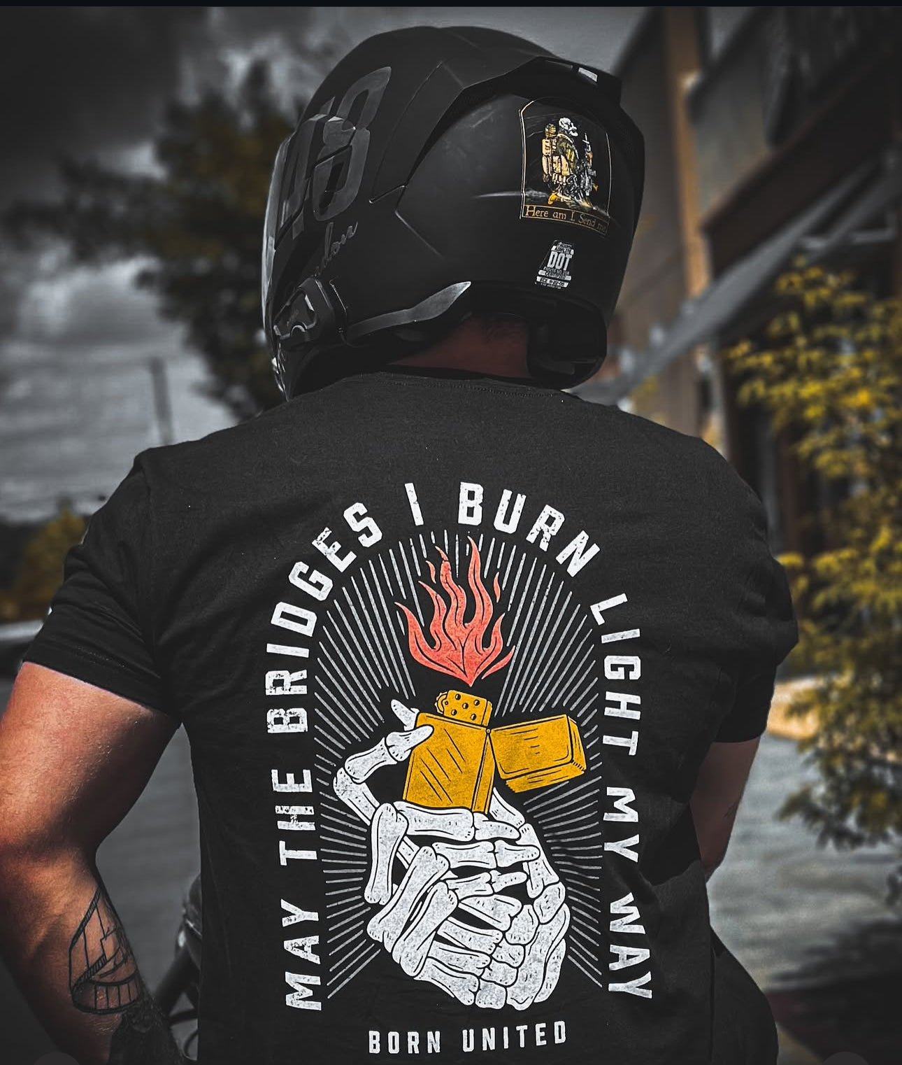 A person wearing a helmet and a black Burning Bridges (Warehouse) tee from Born United Private Label, showcasing a skeleton hand with a flaming lighter. The text reads, MAY THE BRIDGES I BURN LIGHT MY WAY. BORN UNITED, with a building in lush greenery as the backdrop.