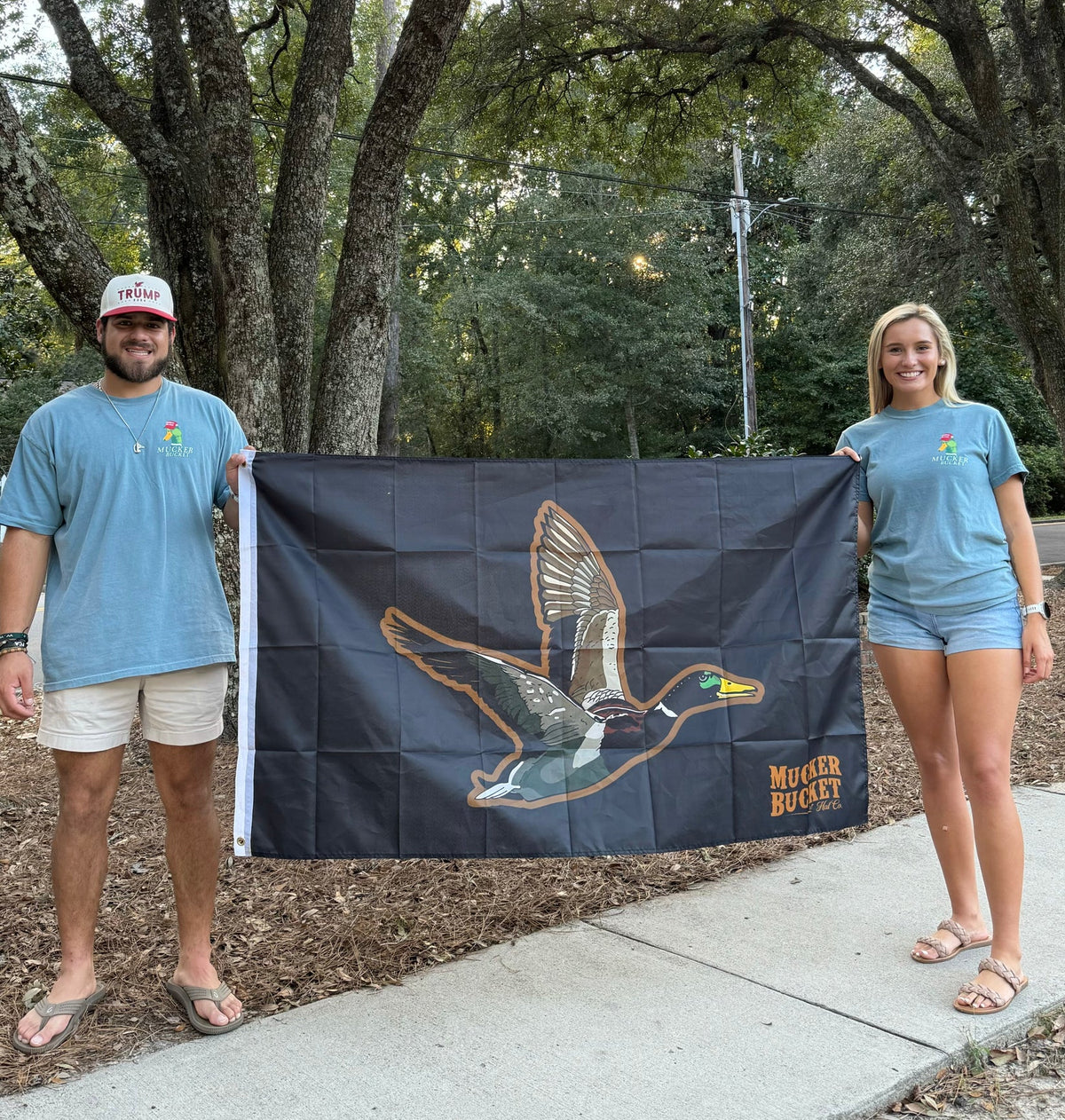 Two people on a tree-lined path hold a Duck It OG flag featuring a flying duck and Mucker Bucket. Theyre wearing blue T-shirts and shorts, embodying the spirit of Duck it.