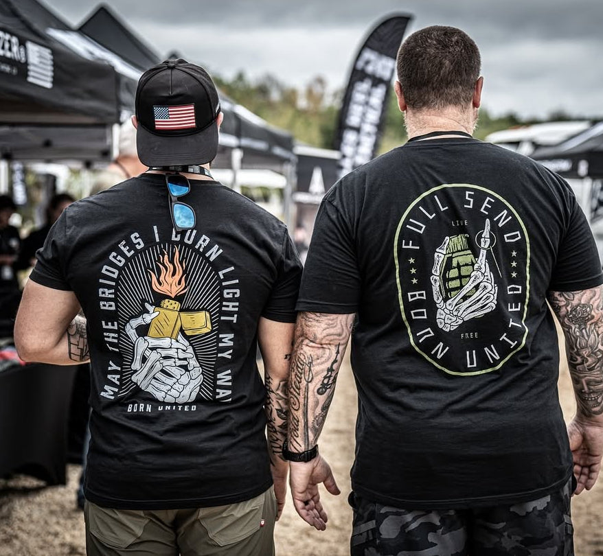 At an outdoor event, two men sport bold black T-shirts. The left shirt reads May the Bridges I Burn Light My Way with subtle flames from Burning Bridges (Warehouse), while the right features Full Send Born United, emphasizing its originality as a Born United Private Label iconic tee.