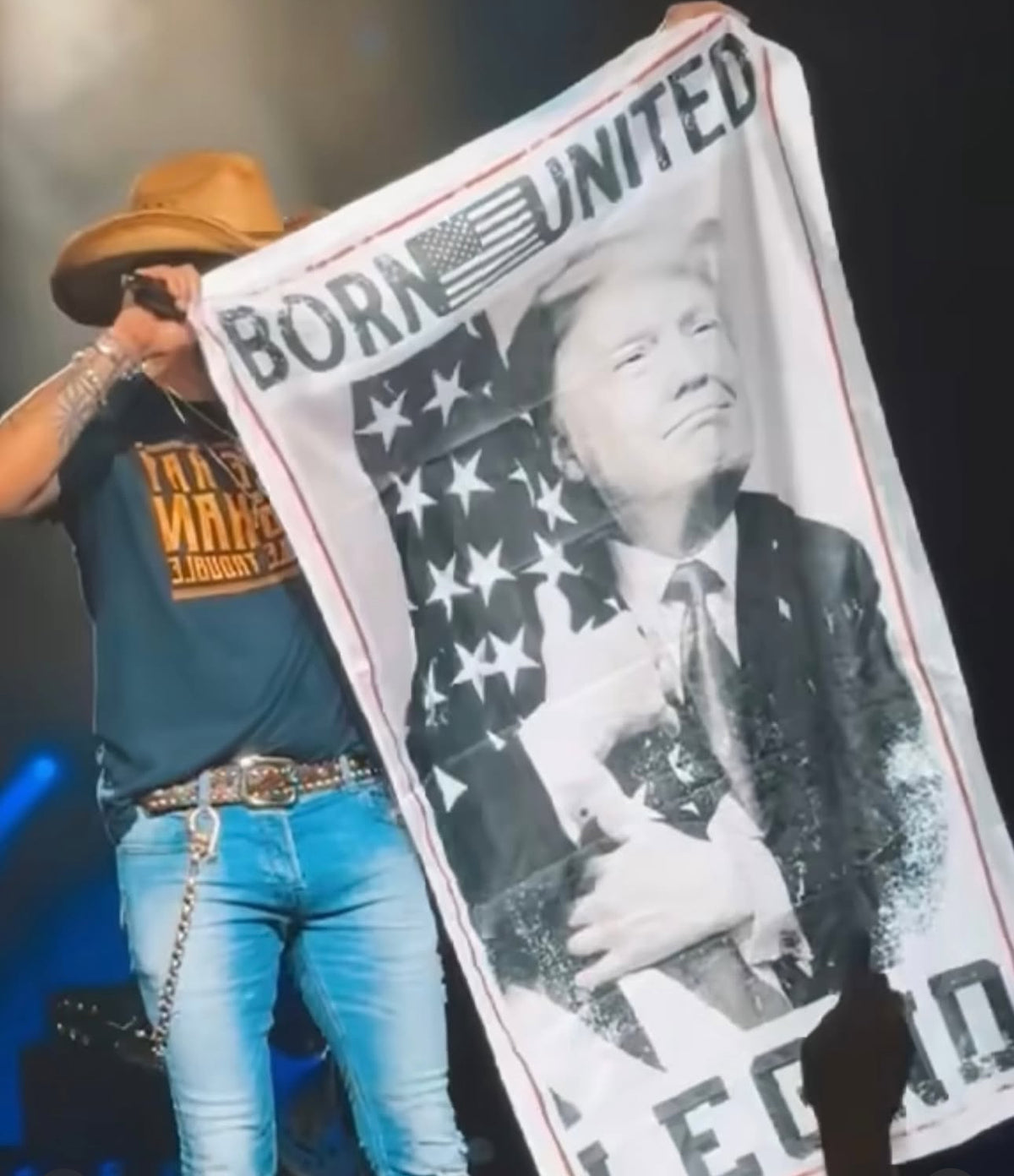 A person in a wide-brimmed hat, jeans, and a T-shirt displays the Legend Trump Flag from Born United Private Label, showcasing President Donald Trump against an American flag with the words Born United, embodying the 2024 election spirit.