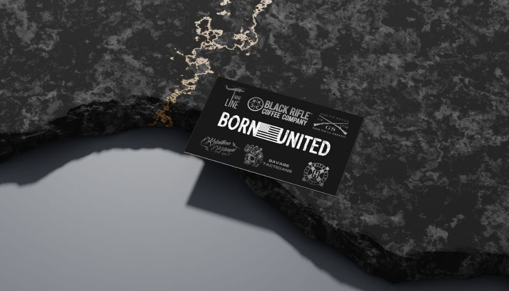 The Born United Gift Card from Gift Cards, featuring the slogan BORN UNITED and various logos, lies on a textured black surface with gold veining, casting a shadow that embodies unity and patriotism.