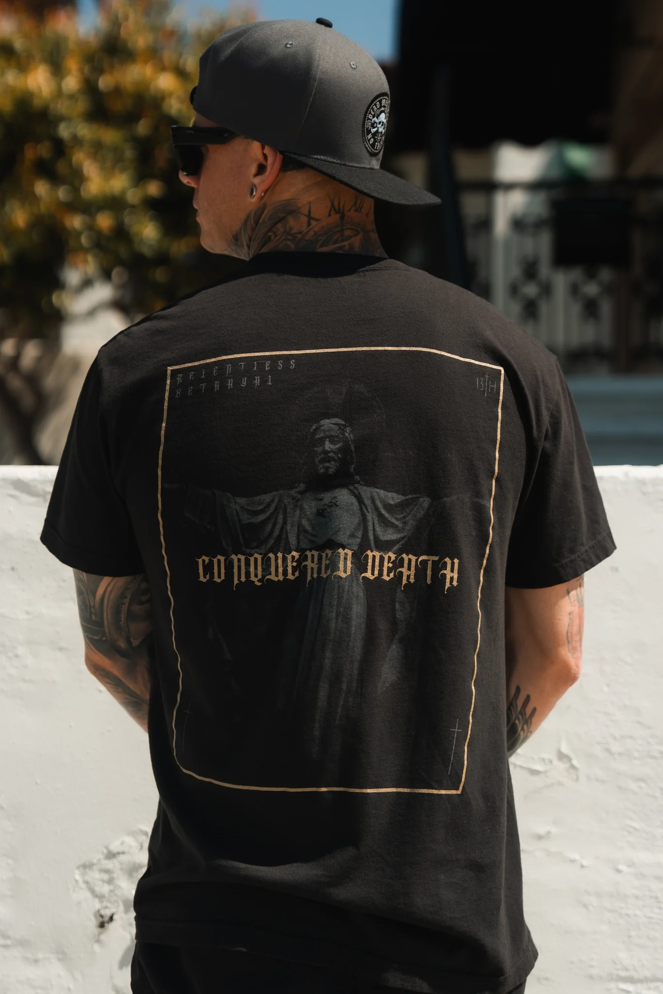 A man in a backwards cap and sunglasses displays the back of a Conquered Death Premium Tee by Relentless Betrayal, featuring a statue image and bold text. He stands against a white wall, with blurred trees and buildings in the background.