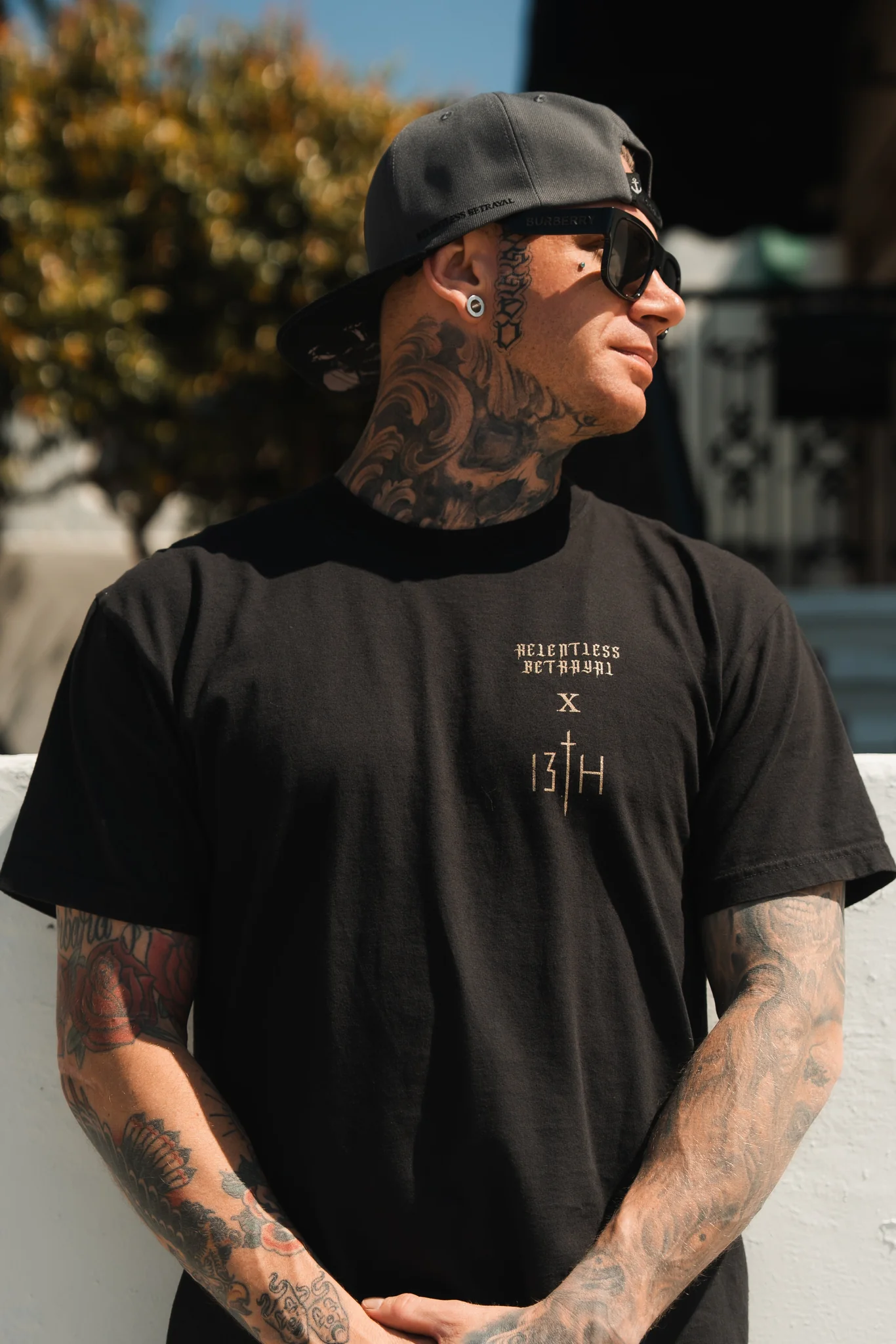 A tattooed person wearing sunglasses and a backwards gray cap is outdoors in a Conquered Death Premium Tee by Relentless Betrayal. Their arms, neck, and head feature intricate tattoos as they look to the side, with a tree and building in the background.