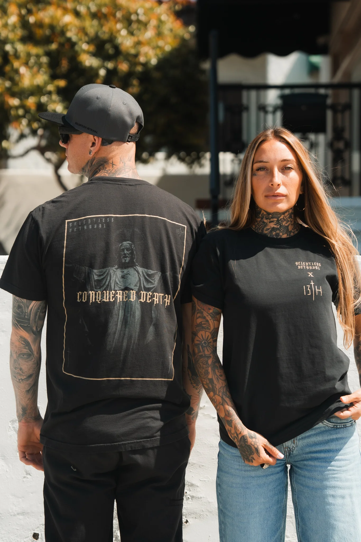 A tattooed man and woman stand outdoors. The man shows off Relentless Betrayals Conquered Death Premium Tee, featuring a statue graphic and the phrase CONQUERED DEATH. The woman faces forward, displaying her arm tattoos. Both sport casual Born United Private Label attire.