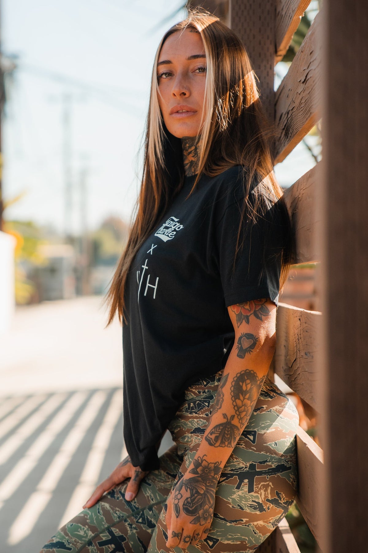 A woman with long brown hair and arm tattoos poses outdoors by a wooden fence in daylight, wearing a black t-shirt and Tango Charlie Apparel's Women's Performance Leggings | Snipes & Stripes, featuring flattering tummy control.