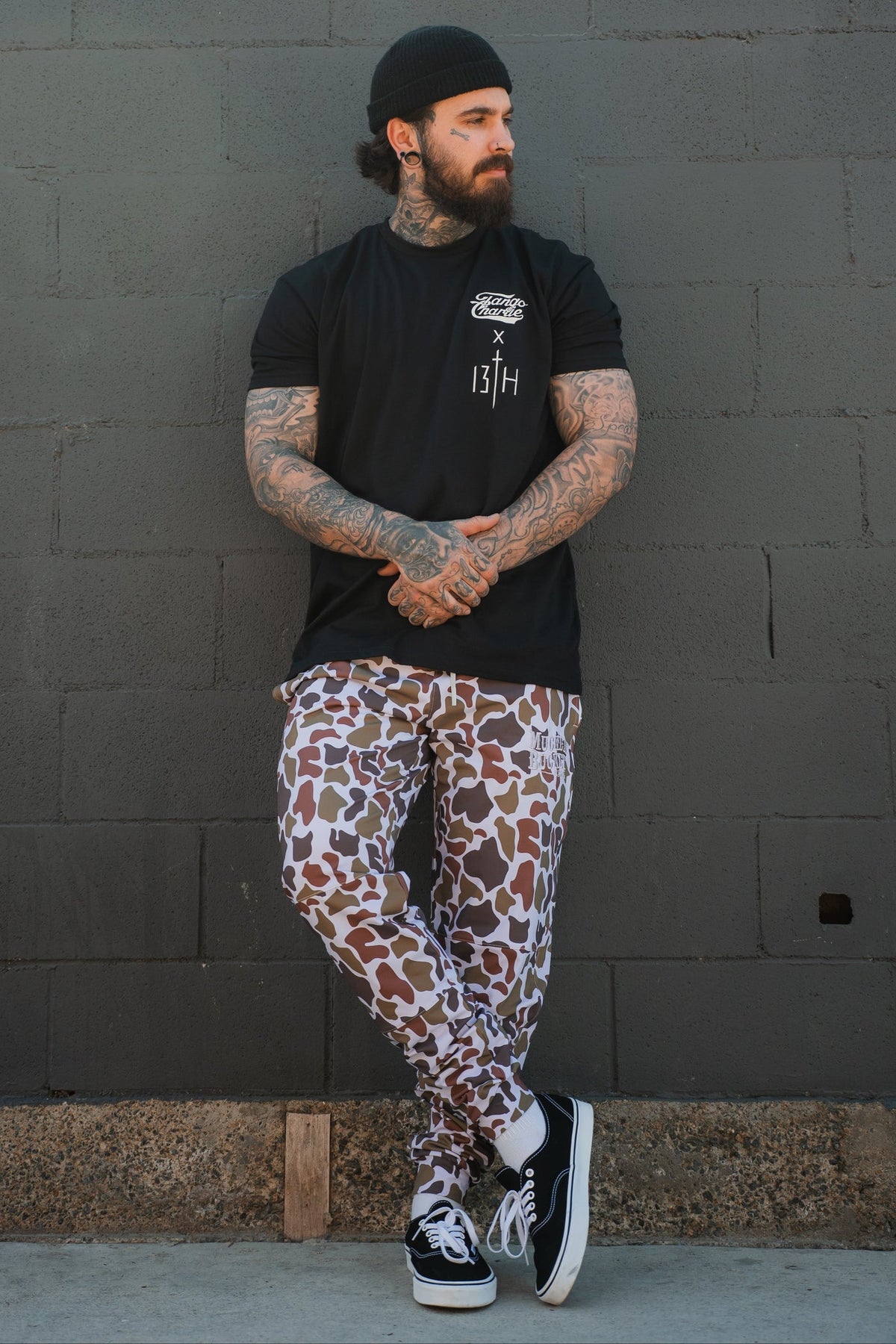 A tattooed, bearded man in a black beanie leans against a gray brick wall wearing a black T-shirt, Mucker Bucket Men's Classic Premium Joggers in Old School Duck Camo, and black Vans sneakers with white soles.