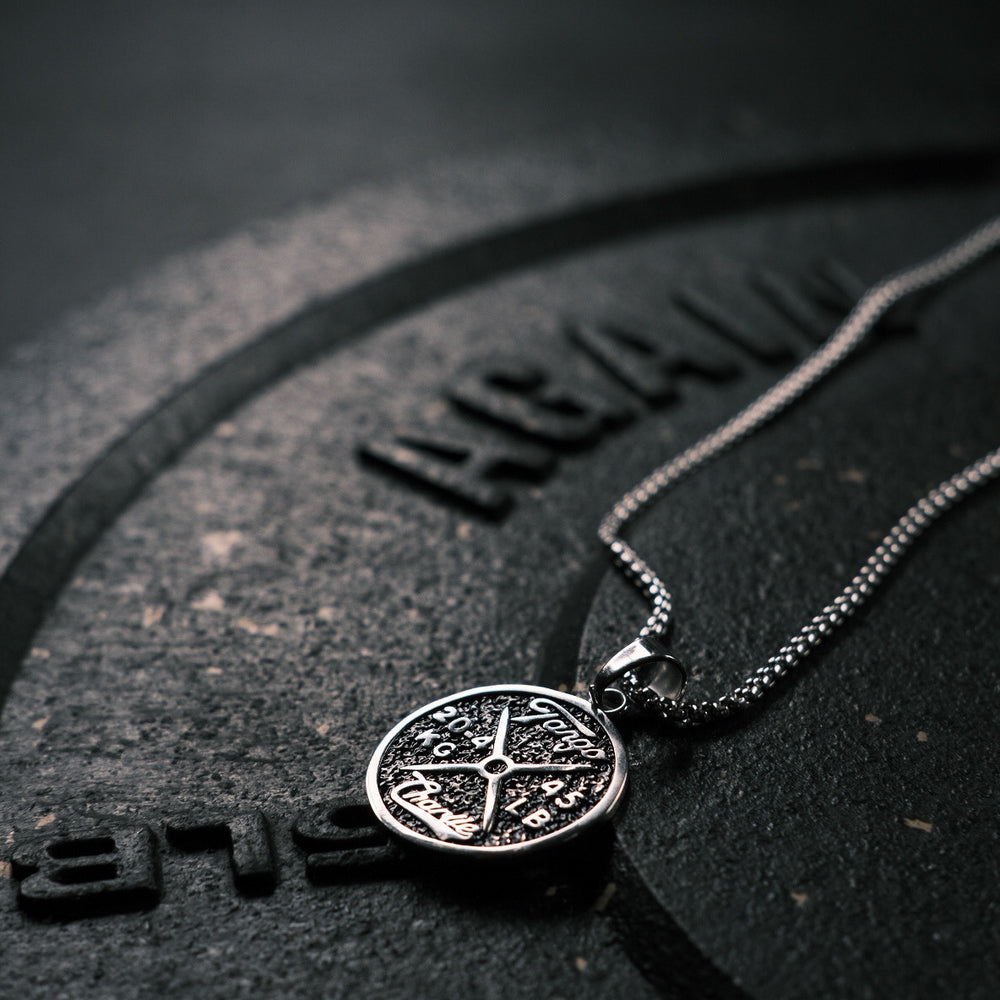A Barbell Necklace by Tango Charlie Apparel lies on a textured dark surface with AGAIN partially visible in embossed letters. The focus is on its elegant and intricate design, combining reliability with a lifetime warranty.