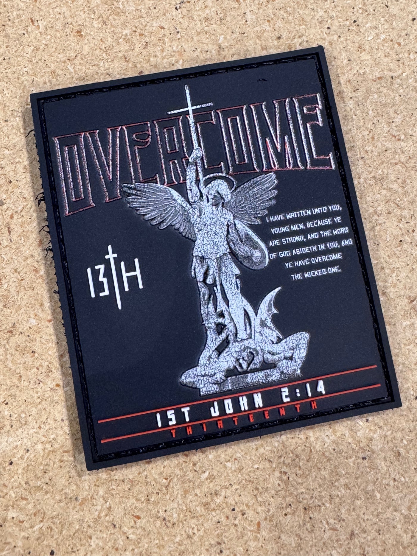 Patch features an armored warrior with wings, sword raised. Text: OVERCOME, 13TH, 1st John 2:14 Thirteenth, and an overcoming evil verse. Black background, silver and red details. Product Name: Overcome PVC Patch by Brand Name: 13th.