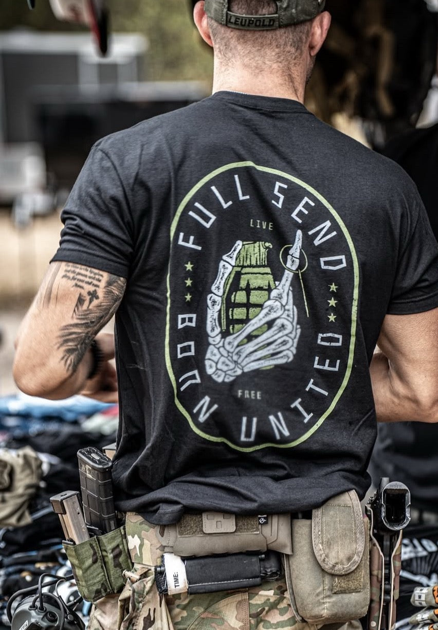 A person in a black Full Send T-shirt by Born United Private Label, featuring a bold skeleton hand design, and camouflage pants stands with their back to the camera. Their belt is loaded with tactical gear like magazine pouches and tools, enhancing the hardcore vibe of their outfit.