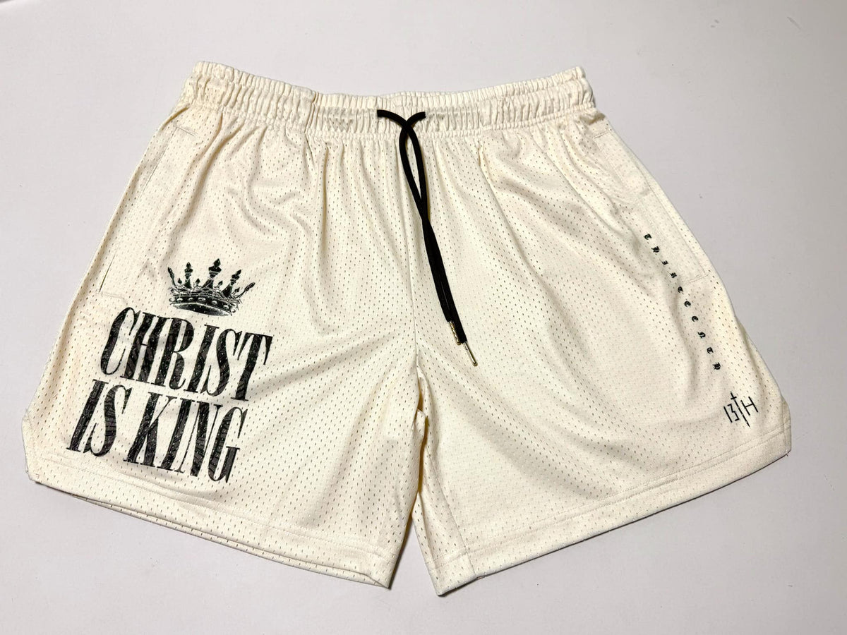 The 13th Men's Mesh Lifestyle Shorts | 5.5" - Christ is King Ivory feature a cream-colored mesh design with "CHRIST IS KING" and a crown graphic printed on the left leg, a black drawstring, and subtle black text on the right leg; these unisex shorts combine comfort and style for everyday wear.