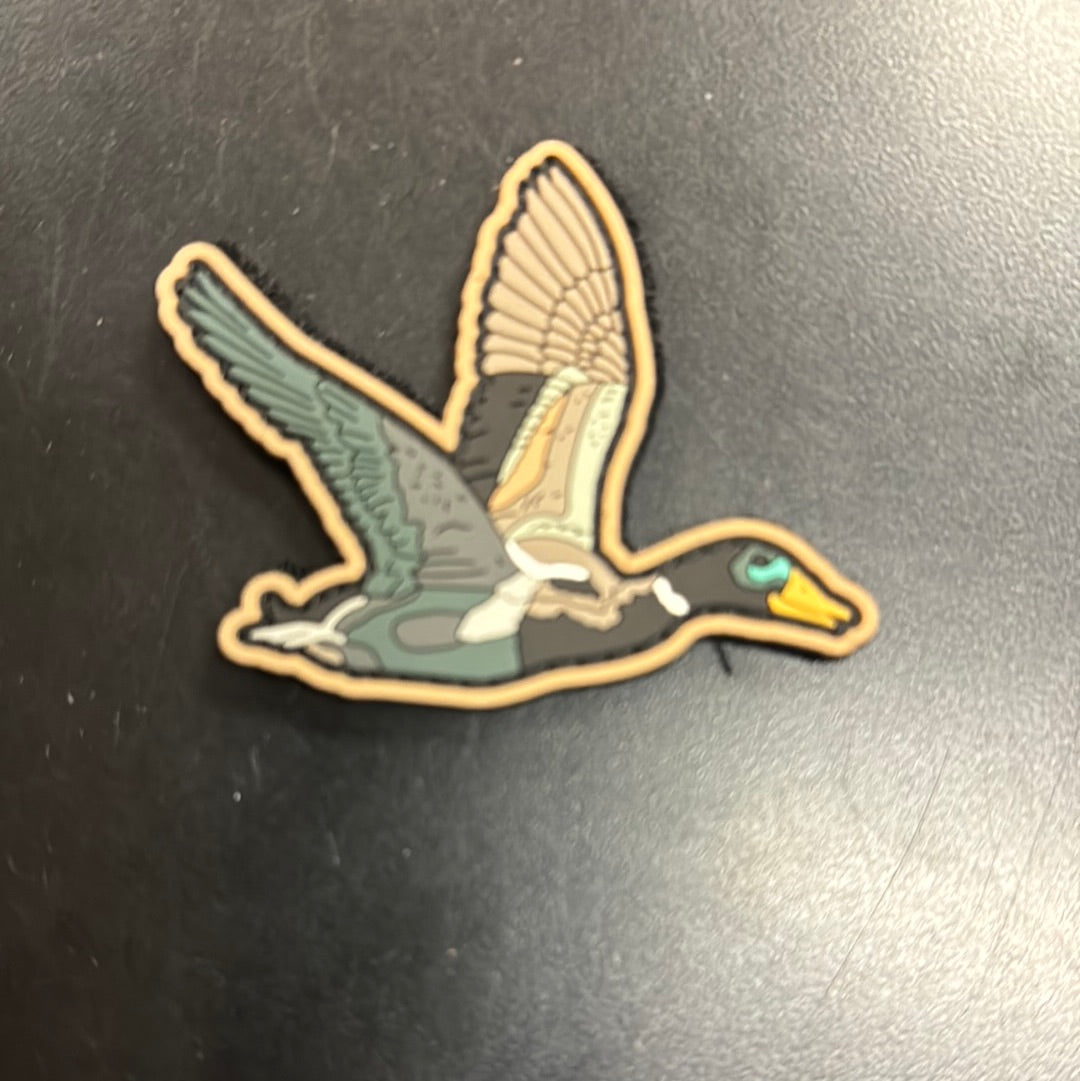 The OG Duck It PVC Patch by Mucker Bucket features a stylized flying duck with detailed wings and a prominent yellow beak, placed on a dark surface. The patch is colorful and eye-catching.