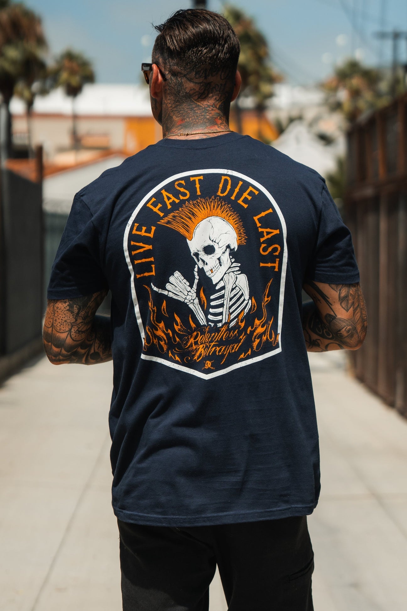 A tattooed individual embodies youth culture, strolling in a Live Fast Navy T-Shirt by Relentless Betrayal. The shirt features a fiery mohawked skeleton with bold text: Live Fast Die Last and Reckless x Drags, capturing an adventurous spirit.