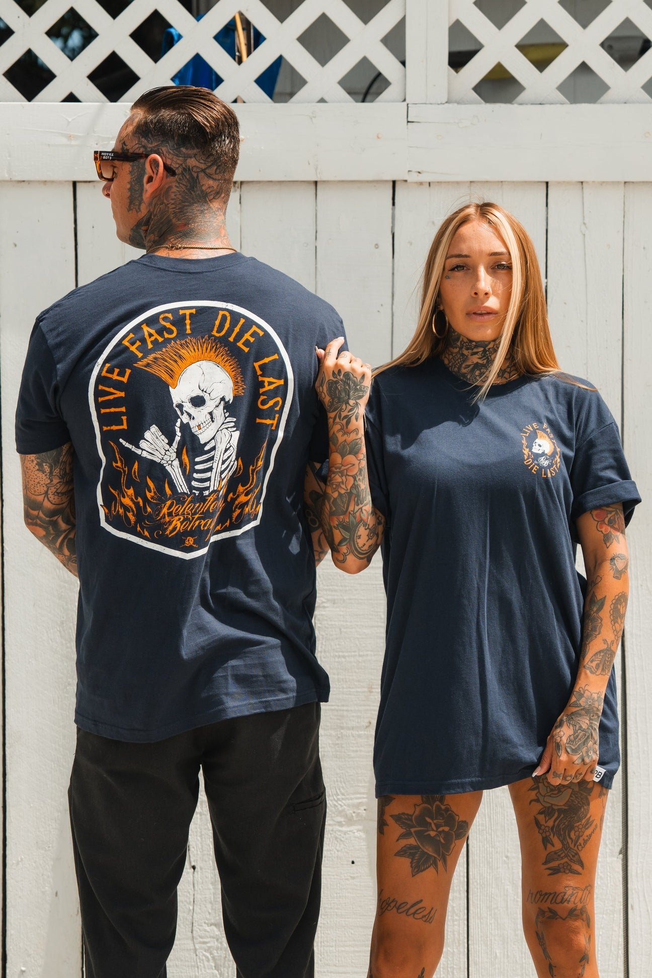 A tattooed man and woman embody youth culture against a white wall, wearing matching Live Fast Navy T-Shirts from Relentless Betrayal. The mans shirt displays a skeleton with Live Fast Die Last on the back, while the womans adorns its front design, capturing a spirit of casual adventure.