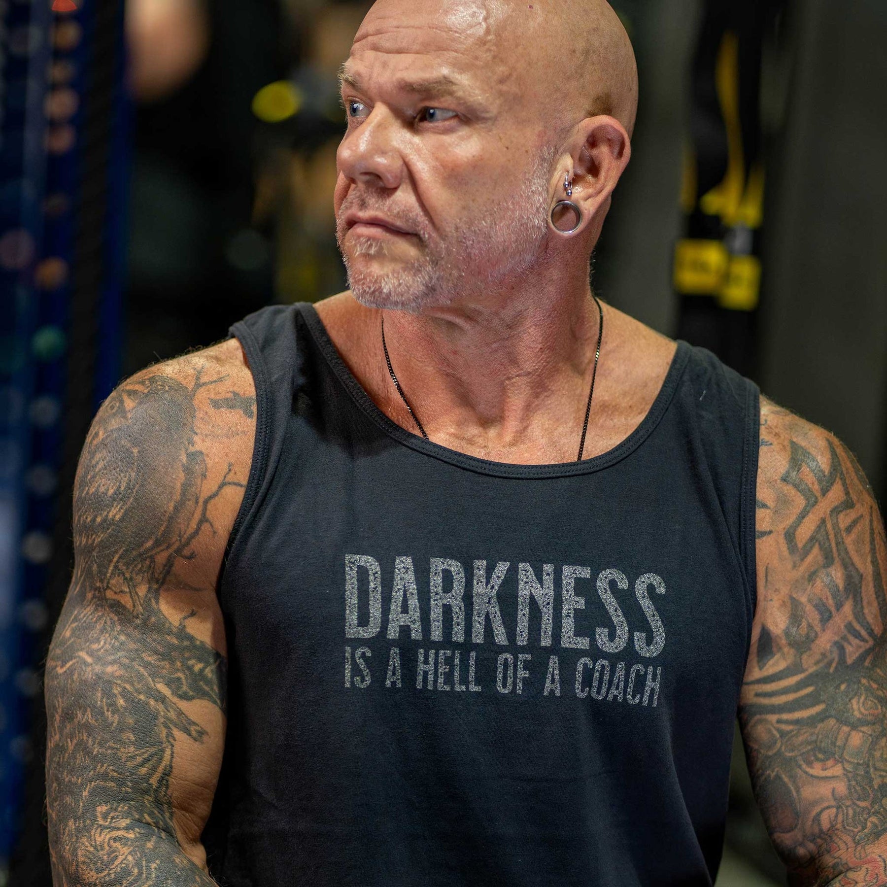 At the gym, a muscular man with tattoos and a shaved head wears a Tango Charlie Apparel Darkness Is A Hell Of A Coach Tank, gazing sideways and embodying triumph over darkness.