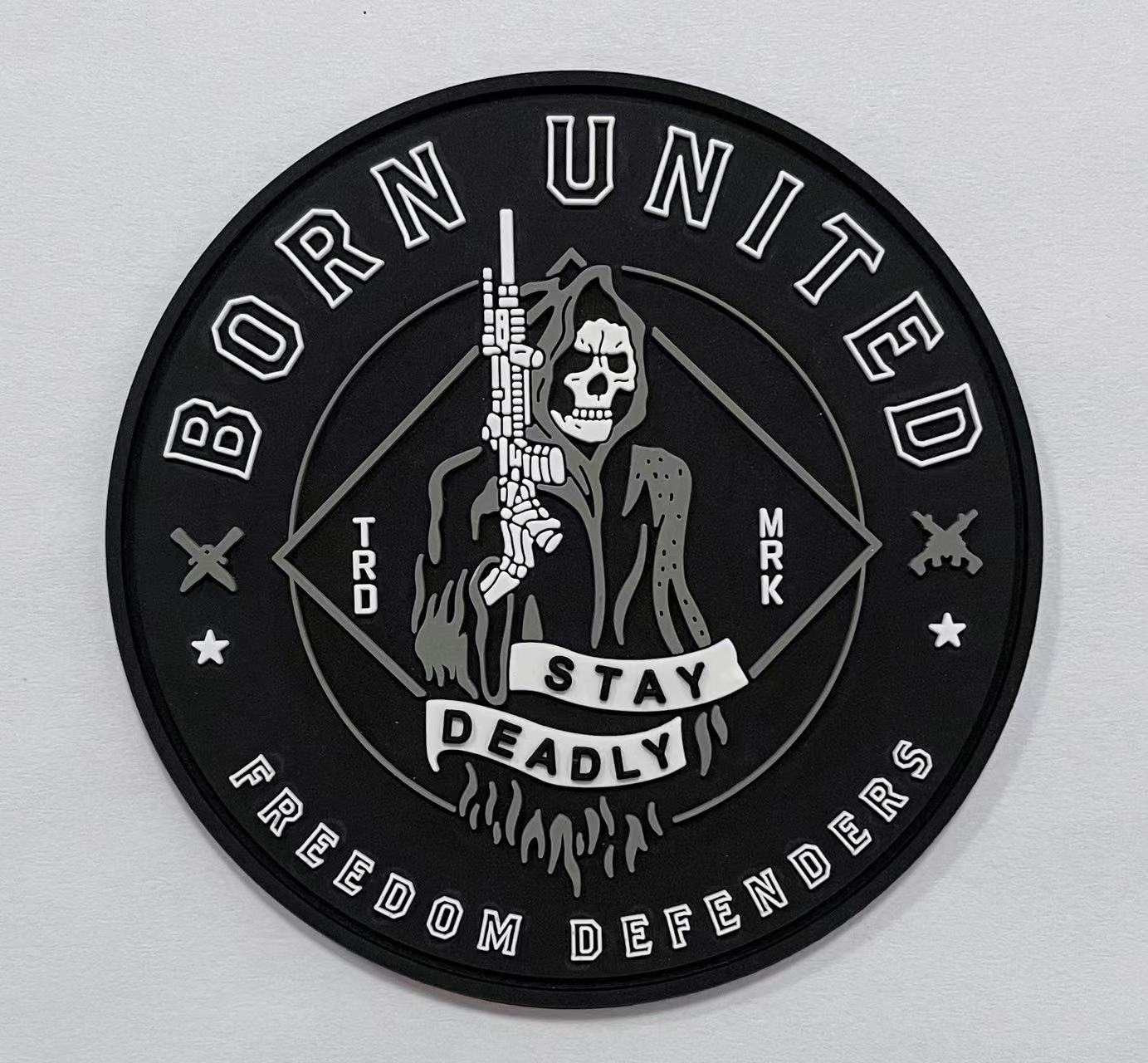 The Stay Deadly PVC Patch by Born United Private Label is a circular black patch featuring a white grim reaper with a rifle, BORN UNITED on top, FREEDOM DEFENDERS on the bottom, and STAY DEADLY on a ribbon across the reaper. Small stars and a diamond shape fill the background.