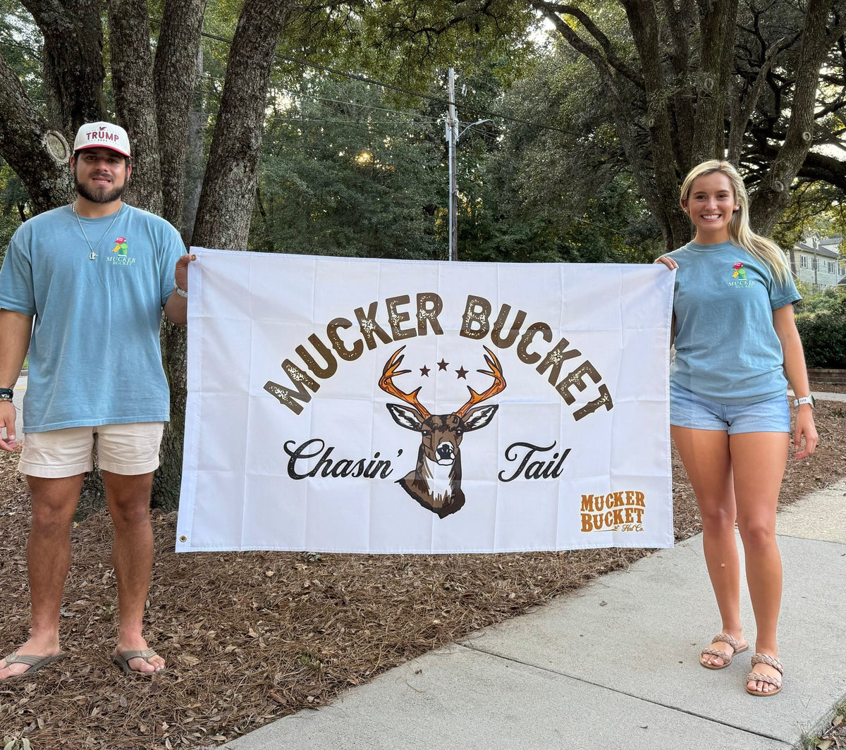 Two people proudly display a Mucker Bucket Chasin’ Tail flag with a deer design, embodying the spirit of outdoor adventure. Clad in blue shirts, they stand on a tree-lined sidewalk, poised for their next journey.