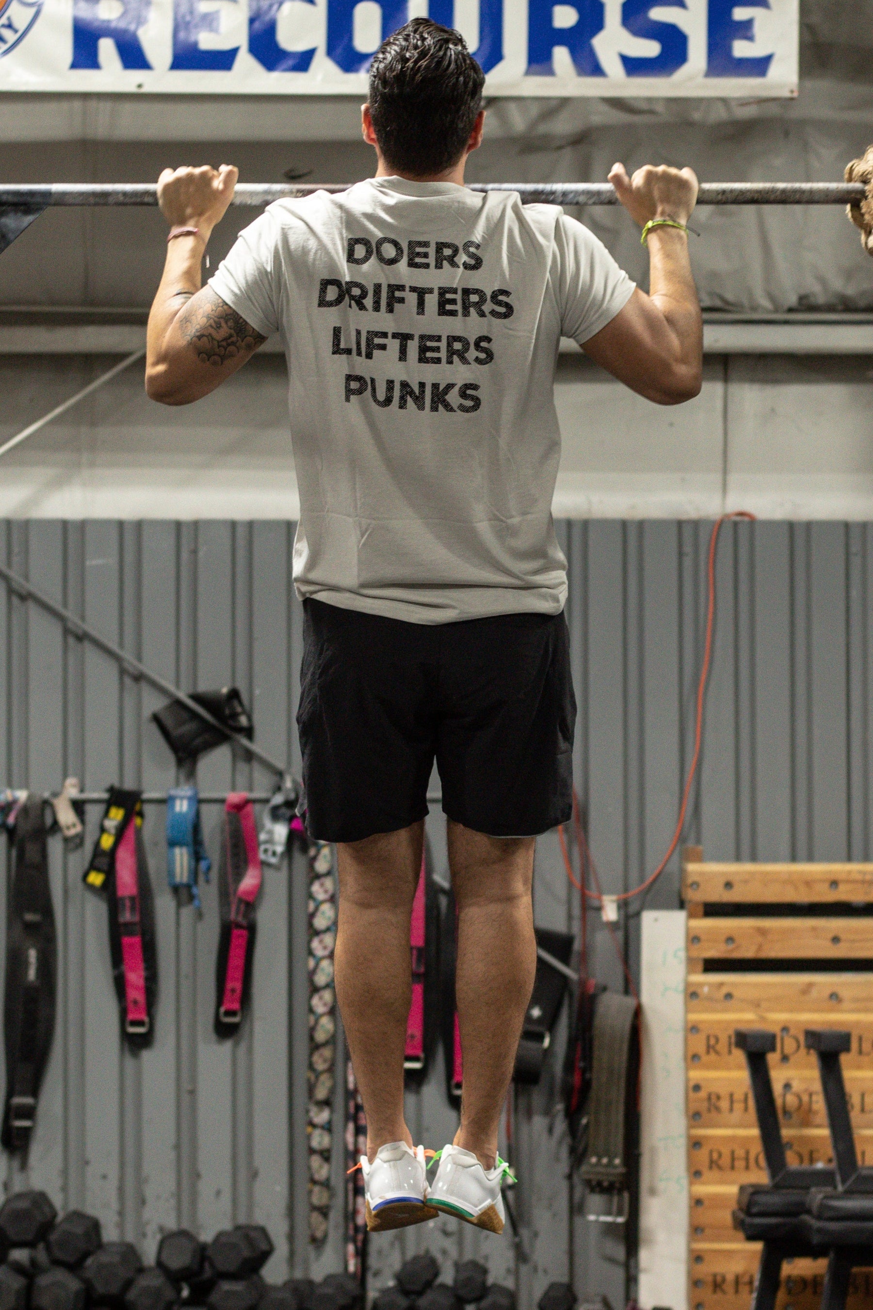 At the gym, a man does a pull-up in the Tango Charlie Apparel Doers Drifters Lifters Punks Unisex Tee. Surrounded by fitness gear, he reps your crew in style with this bold back-print shirt.