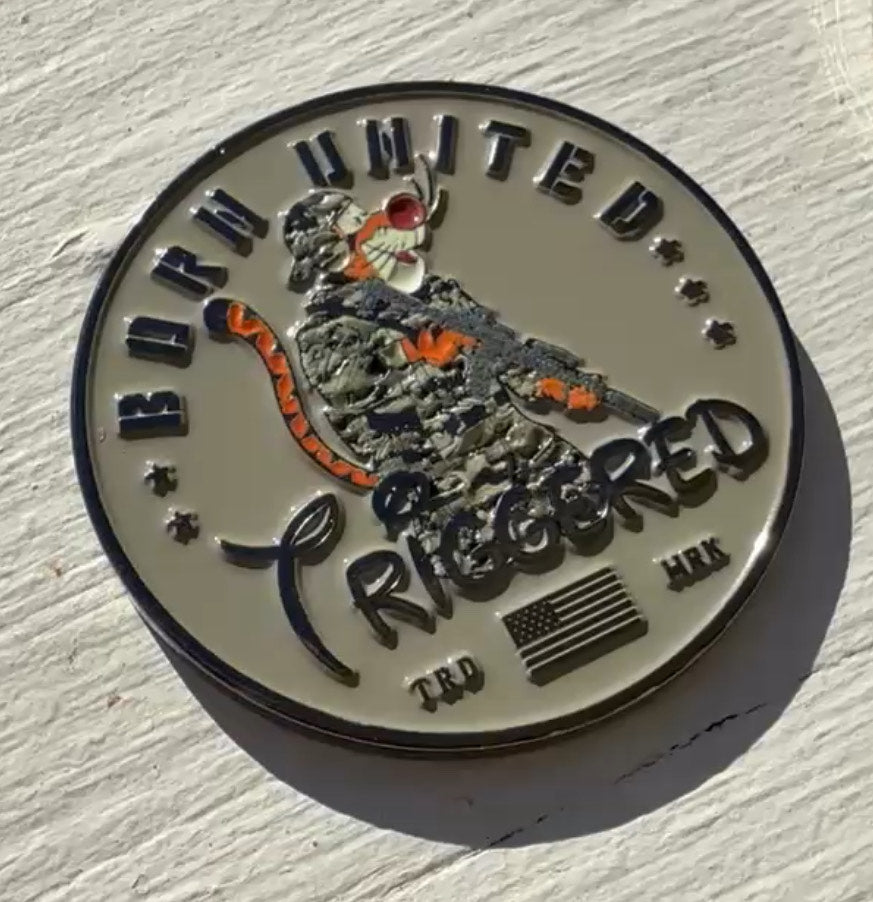 The Triggered Challenge Coin by Born United Private Label displays Both United above and Triggered below an armed tiger in a helmet with goggles. An American flag appears at the bottom next to TRD MRK, all textured by shadows on this round metal piece.