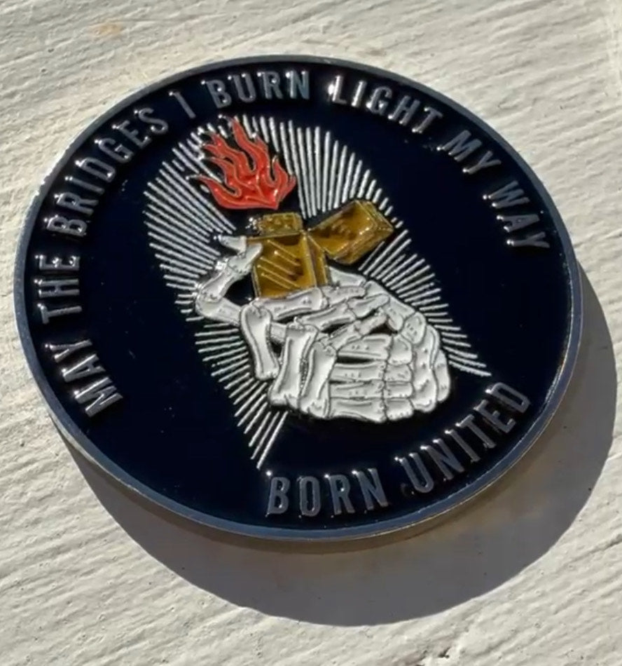 The Burning Bridges Challenge Coin by Born United Private Label displays a skeleton hand with a flaming match. Around the design are the words MAY THE BRIDGES I BURN LIGHT MY WAY atop and BORN UNITED at the bottom, set on a sunlit, light-colored background.