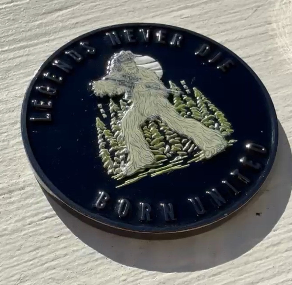 The Legends Never Die Challenge Coin by Born United Private Label is a black coin featuring a silver Bigfoot in a forest with the moon behind. LEGENDS NEVER DIE and BORN UNITED are engraved on the border, capturing mystery and unity.