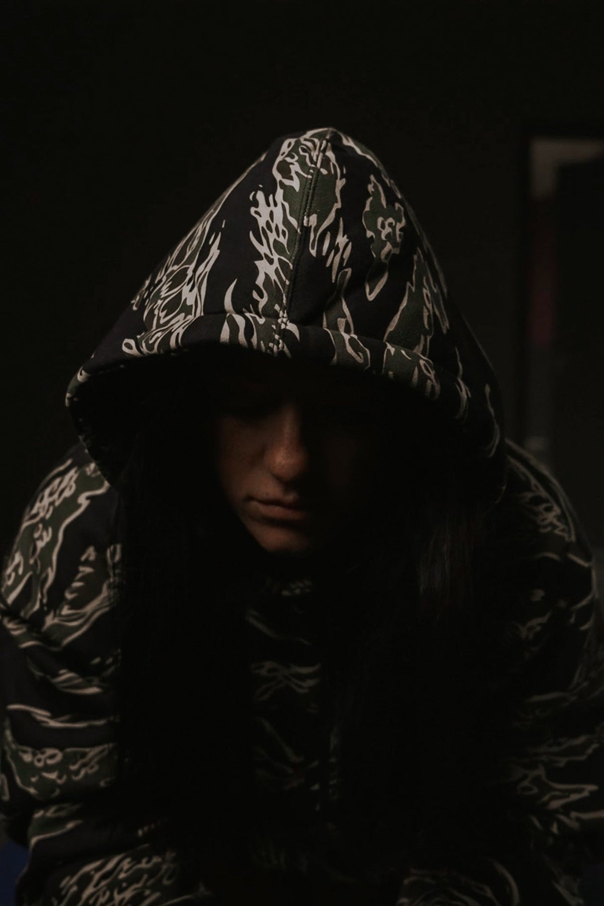 A person wearing the "No One Is Coming To Save You" Premium Hoodie by Tango Charlie Apparel sits in a dimly lit setting, their face partly hidden by the hood, evoking a mysterious sense of self-motivation and responsibility.