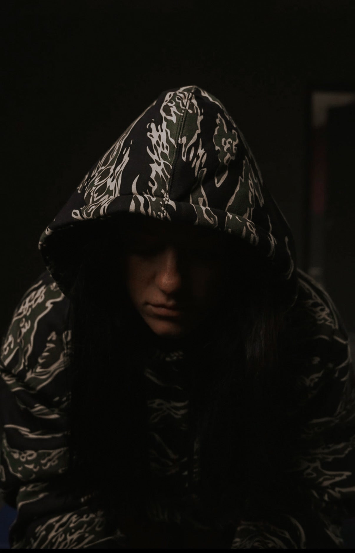 In a dimly lit space, someone wears the No One Is Coming To Save You hoodie by Tango Charlie Apparel. The hood shades their face, enhancing the dark backgrounds mysterious and intense atmosphere, hinting at their journey of self-reliance and pursuit of life goals.
