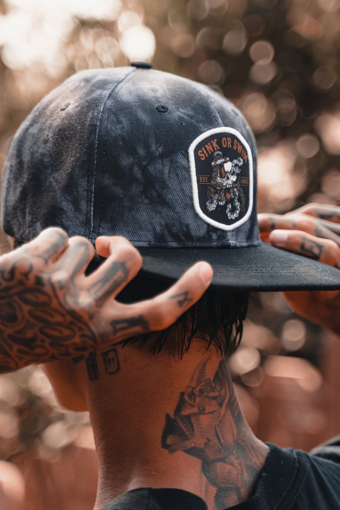 A tattooed individual adjusts a Relentless Betrayal Sink Or Swim Tie Dye snapback in a warmly lit setting, emphasizing tattoos on their neck and arms.