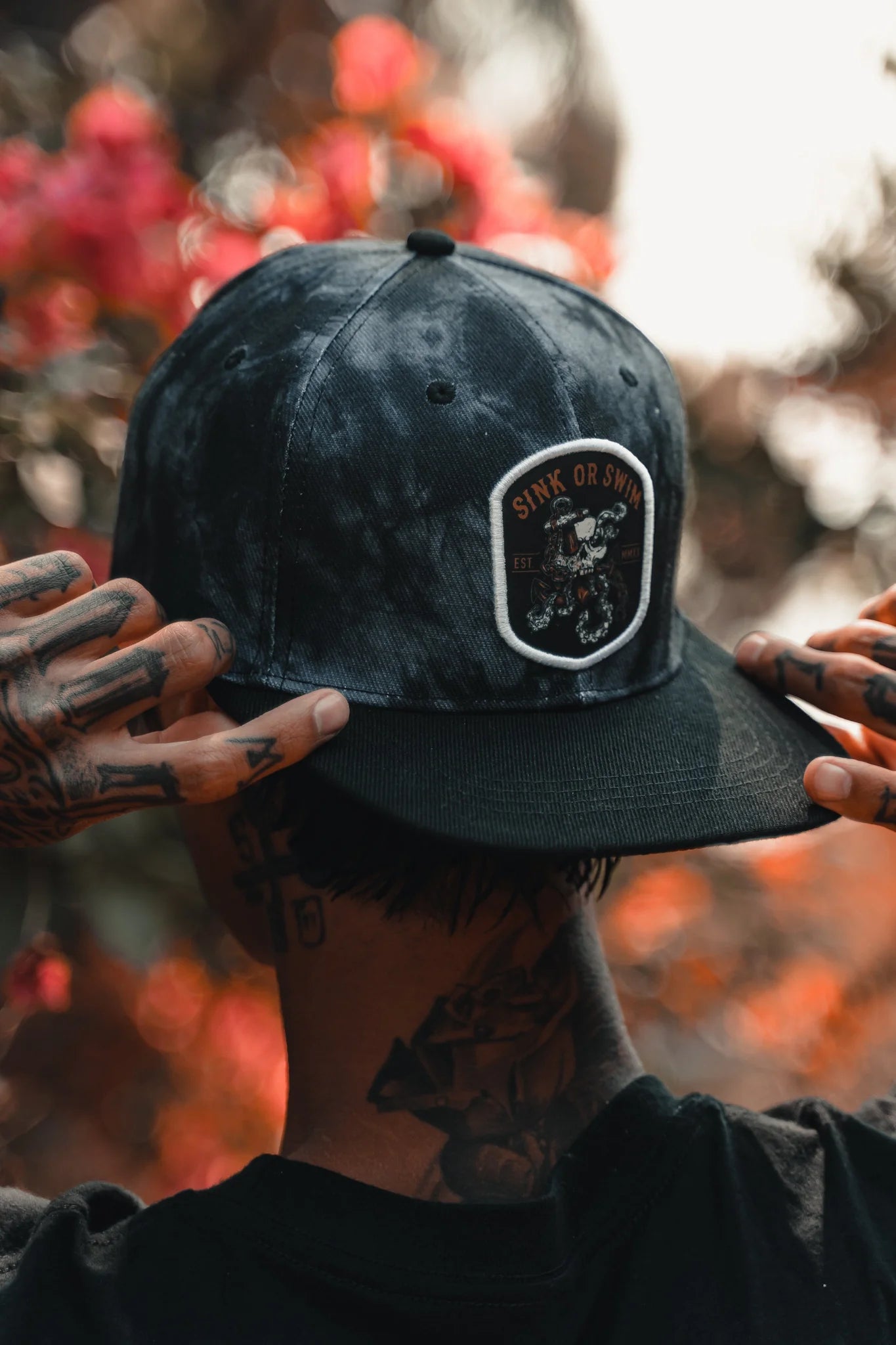 A tattooed individual adjusts a Sink Or Swim Tie Dye snapback from Relentless Betrayal, featuring a skull patch. The background is an artful blur of pink flowers and greenery.