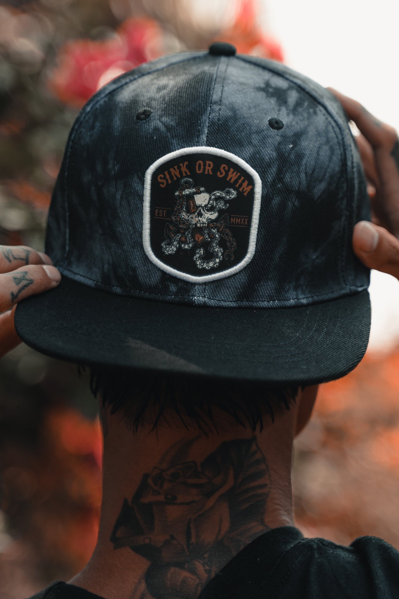 A person dons a Relentless Betrayal Sink Or Swim Tie Dye dark baseball cap. Tattoos decorate their hands and neck, accentuating their edgy style against a blurred background with red and green hues.