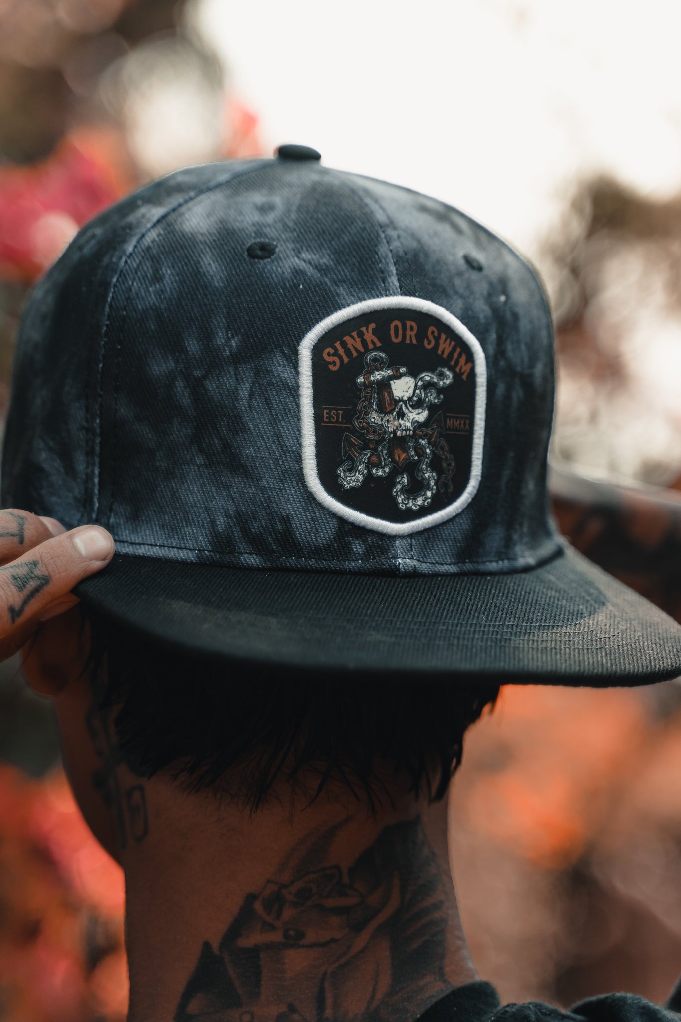A person facing away adjusts a Relentless Betrayal Sink Or Swim Tie Dye snapback, featuring an octopus patch. Their tattooed hands and neck stand out against a blurred background with warm tones.