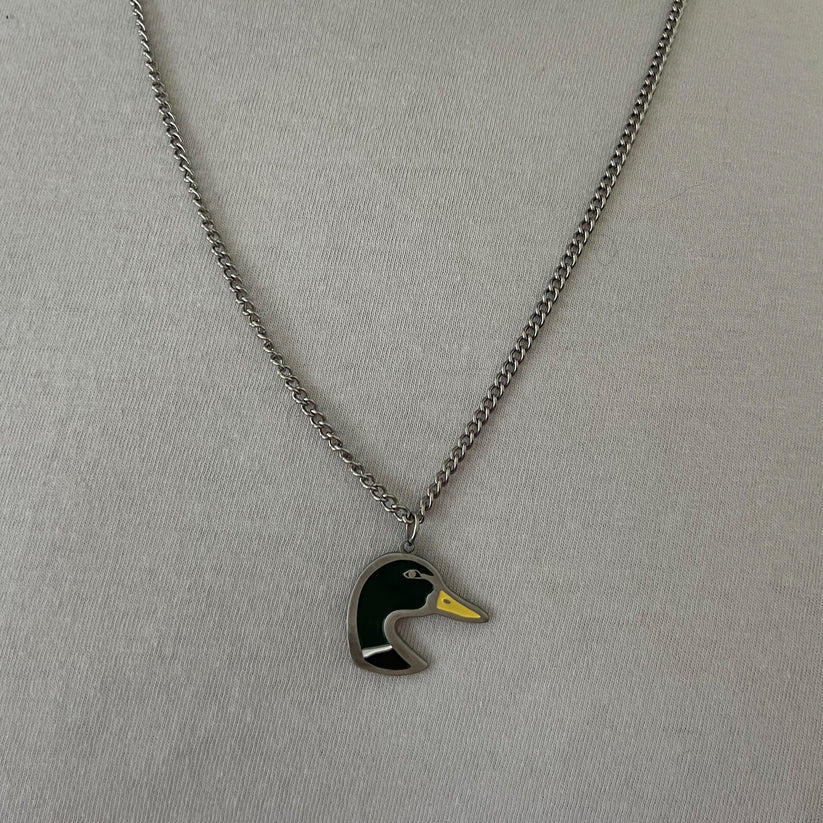 The Mallard OG Stainless Steel Necklace by Mucker Bucket features a pendant of a stylized duck head in green, yellow, and black. Made in the USA for quality craftsmanship, it is displayed on a silver chain against a light gray background.
