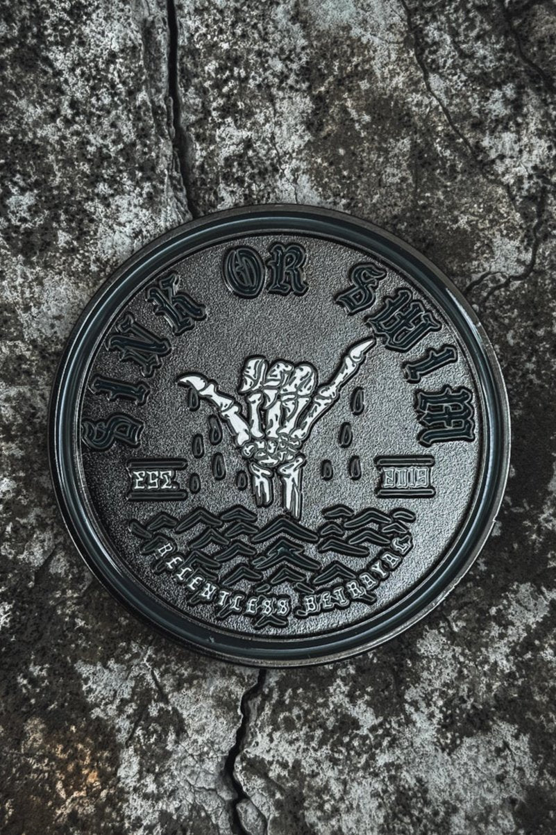 The Sink Or Swim Challenge Coin by Relentless Betrayal is a round stainless steel coin that rests on a rough, cracked surface. It features a raised hand in the shaka sign surrounded by waves, with Sink or Swim and Fearless Summer embossed around the edge, symbolizing resilience and perseverance.