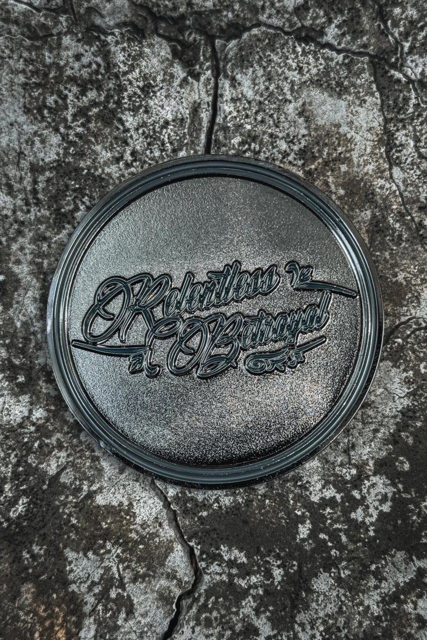 The Sink Or Swim Challenge Coin by Relentless Betrayal is a round, textured black coin with intricate cursive on top. It rests on a rough, cracked stone surface. A glare partially obscures the ornate text, enhancing its vintage feel of perseverance.