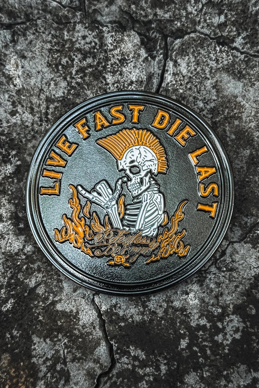 The Relentless Betrayals Live Fast Die Last Challenge Coin features a handcrafted stainless steel emblem with a skeleton sporting a mohawk and holding a cigarette amid flames. It bears the text LIVE FAST DIE LAST above and Rebel At Heart below, set against a cracked stone background.
