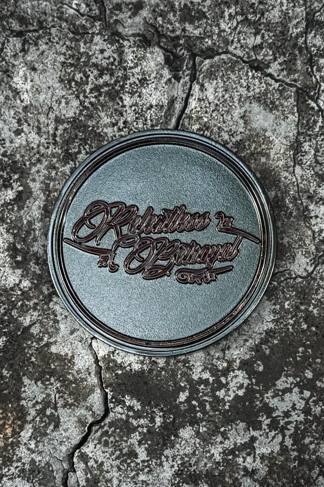 The All Gas No Brakes Challenge Coin by Relentless Betrayal epitomizes style, like a round, dark green coaster with ornate brown text against cracked gray concrete.
