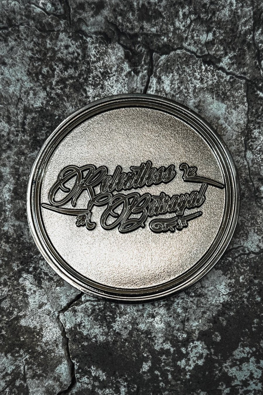 The Vengeance Challenge Coin by Relentless Betrayal, an ornate round metallic plaque with Rebels of Baroque engraved and decorative flourishes, lies on a cracked stone surface. Its shiny finish contrasts strikingly against the textured background, celebrating resilience.