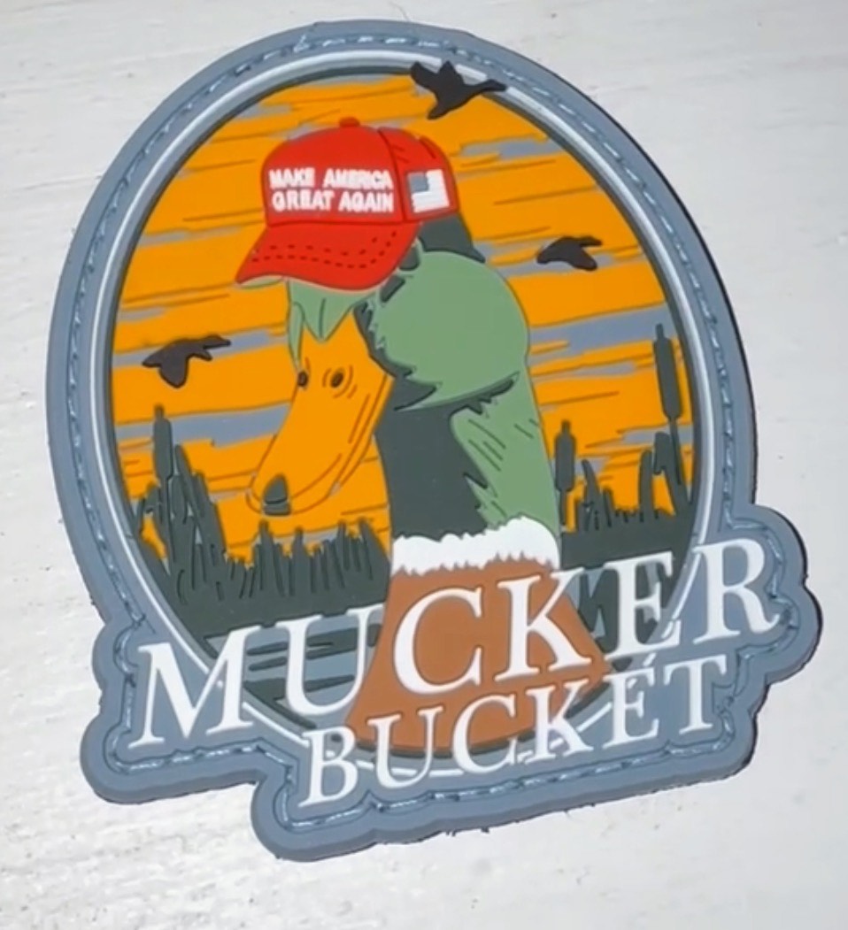 The Smart Duck PVC Patch by Mucker Bucket features a clever duck in a Make America Great Again hat against an orange sunset, silhouetted with cacti and birds.