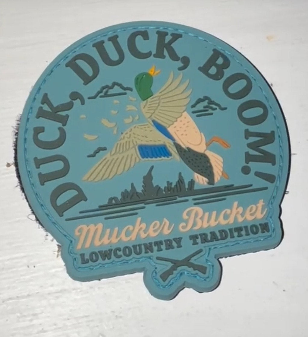 The Duck Duck Boom PVC Patch by Mucker Bucket shows a duck flying over grass with the text DUCK, DUCK, BOOM! A light blue background complements the illustration, with Mucker Bucket and LOWCOUNTRY TRADITION in varied fonts.
