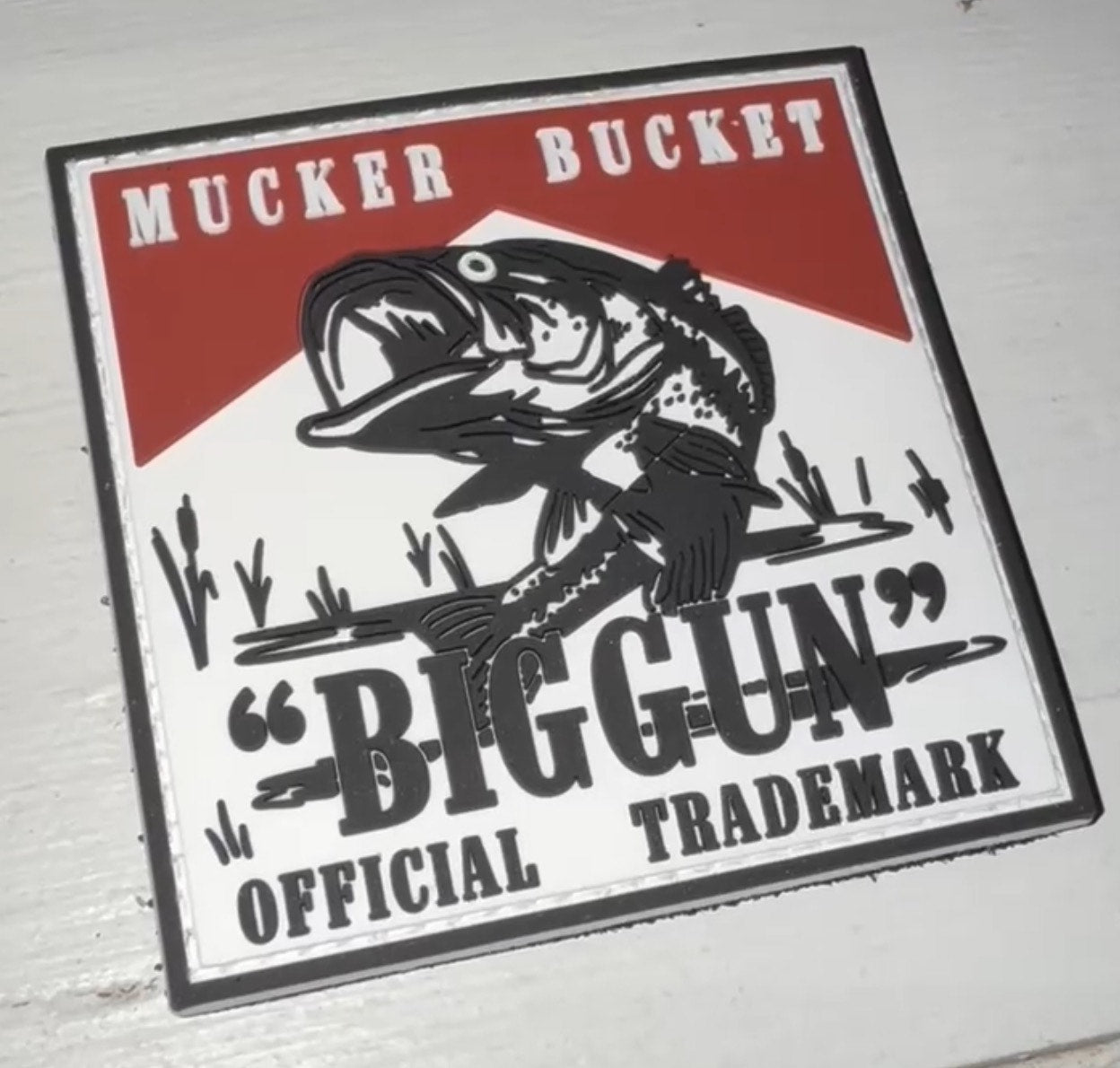 Sticker featuring Mucker Buckets Biggun PVC Patch logo—an illustrated fish emerging from a barrel set against a bold red and white background, with the text Mucker Bucket Biggun Official Trademark.